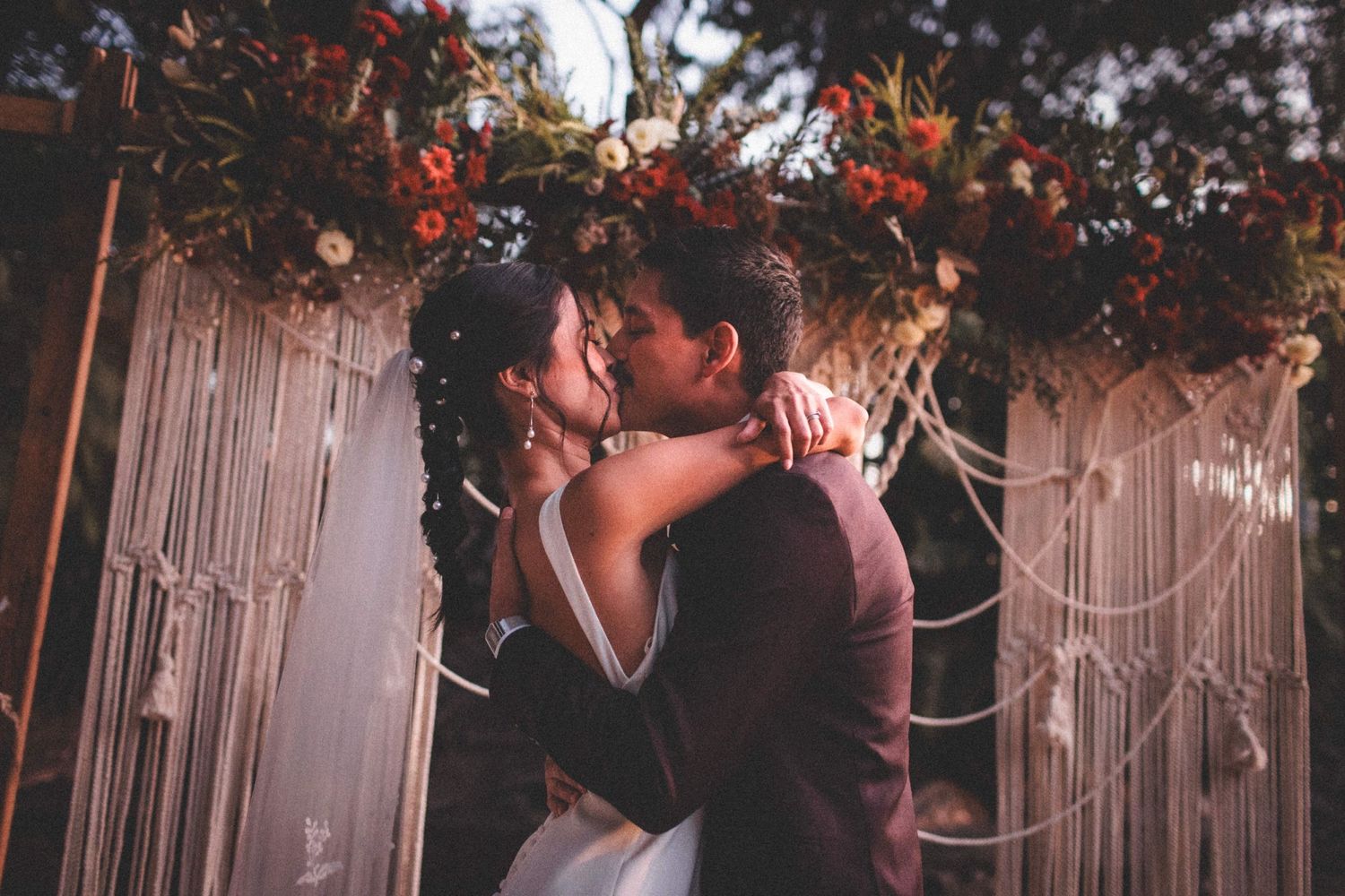 A romantic wedding couple embraces under a macrame backdrop decorated with autumn florals and twinkling lights.