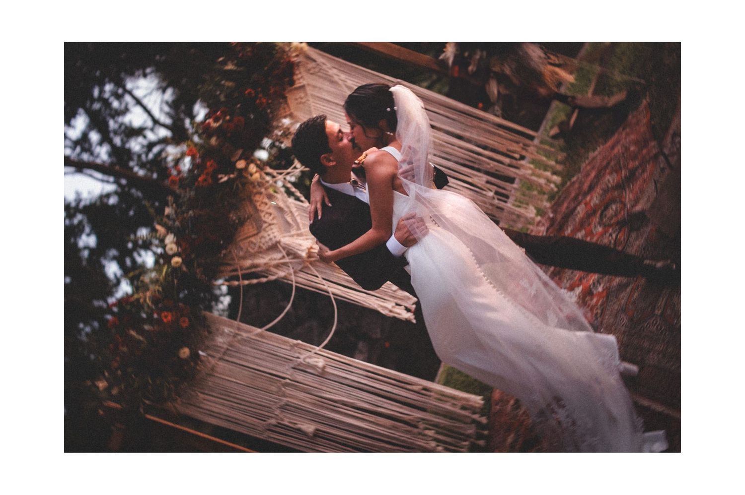A romantic couple in wedding attire sits together on a rustic outdoor wooden swing during sunset.