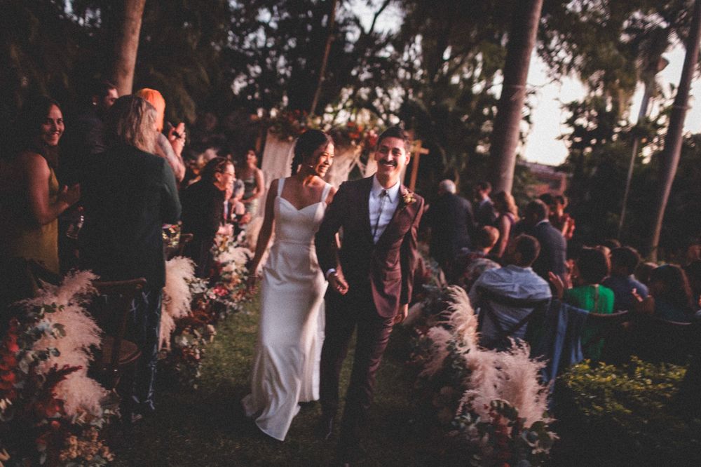 A celebratory wedding party dances outdoors at dusk among tall trees and decorative pampas grass.