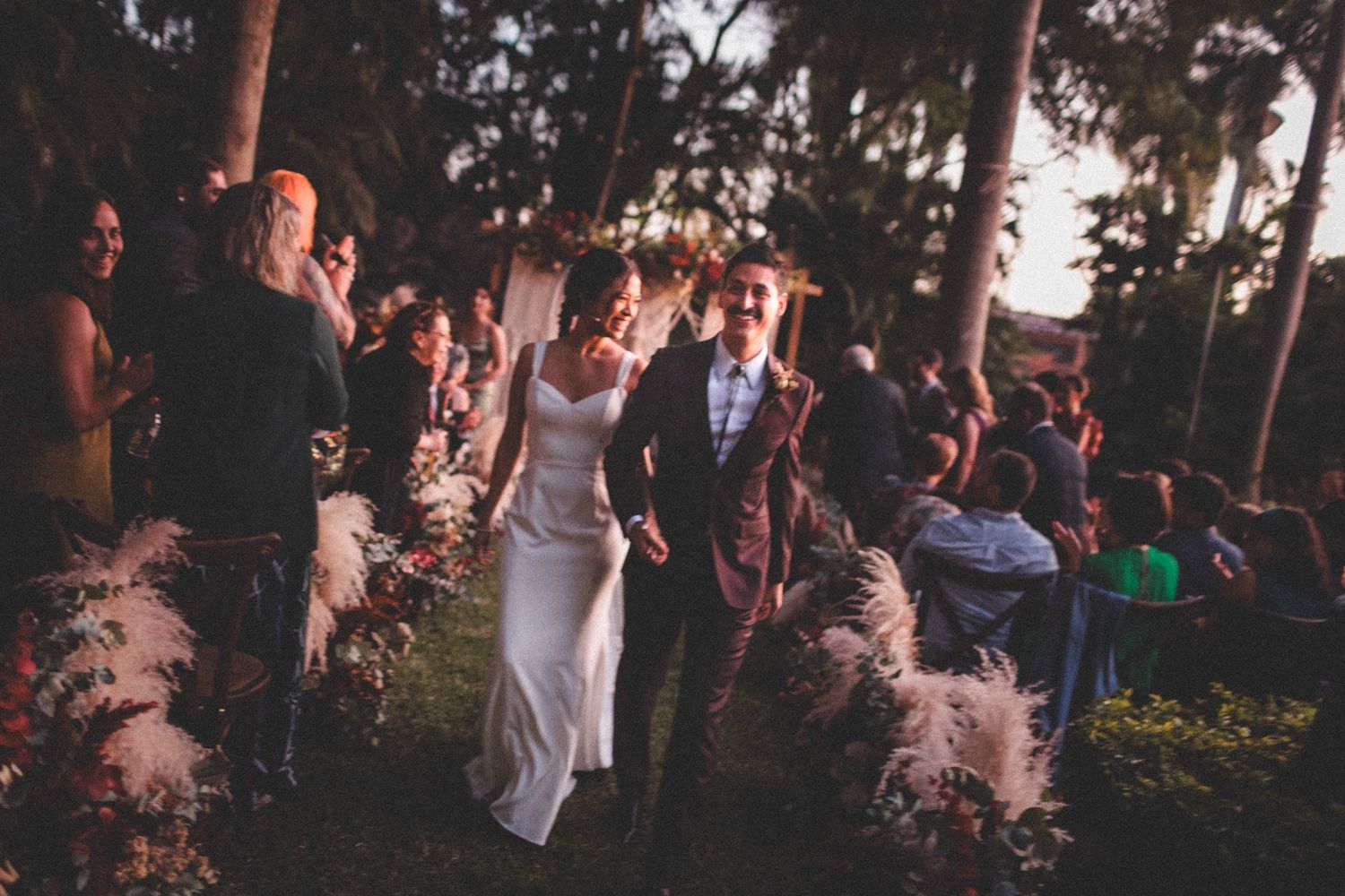 A celebratory wedding party dances outdoors at dusk among tall trees and decorative pampas grass.