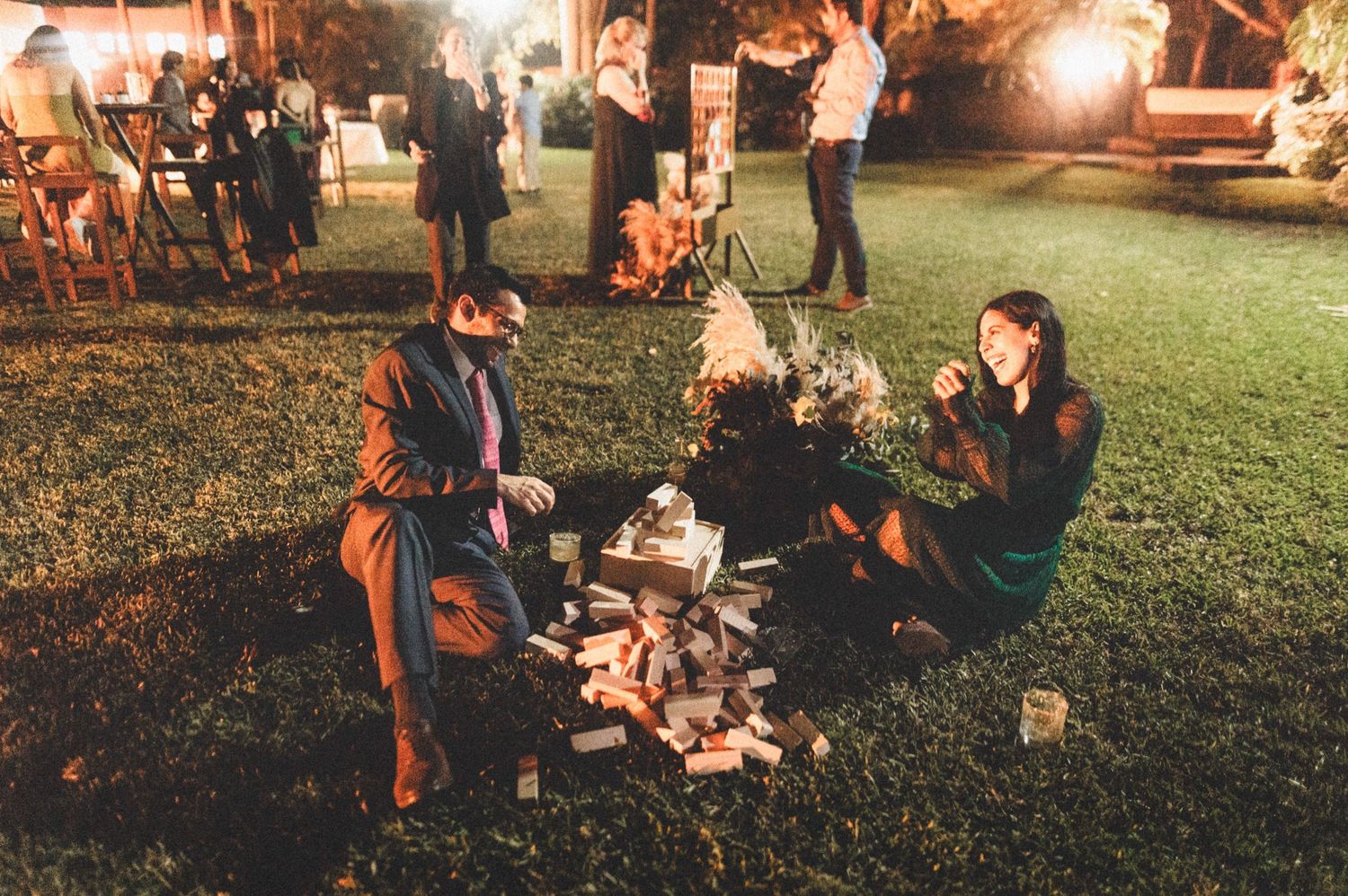 People gather outdoors on a lawn for an evening celebration with warm lighting and casual seating arrangements.