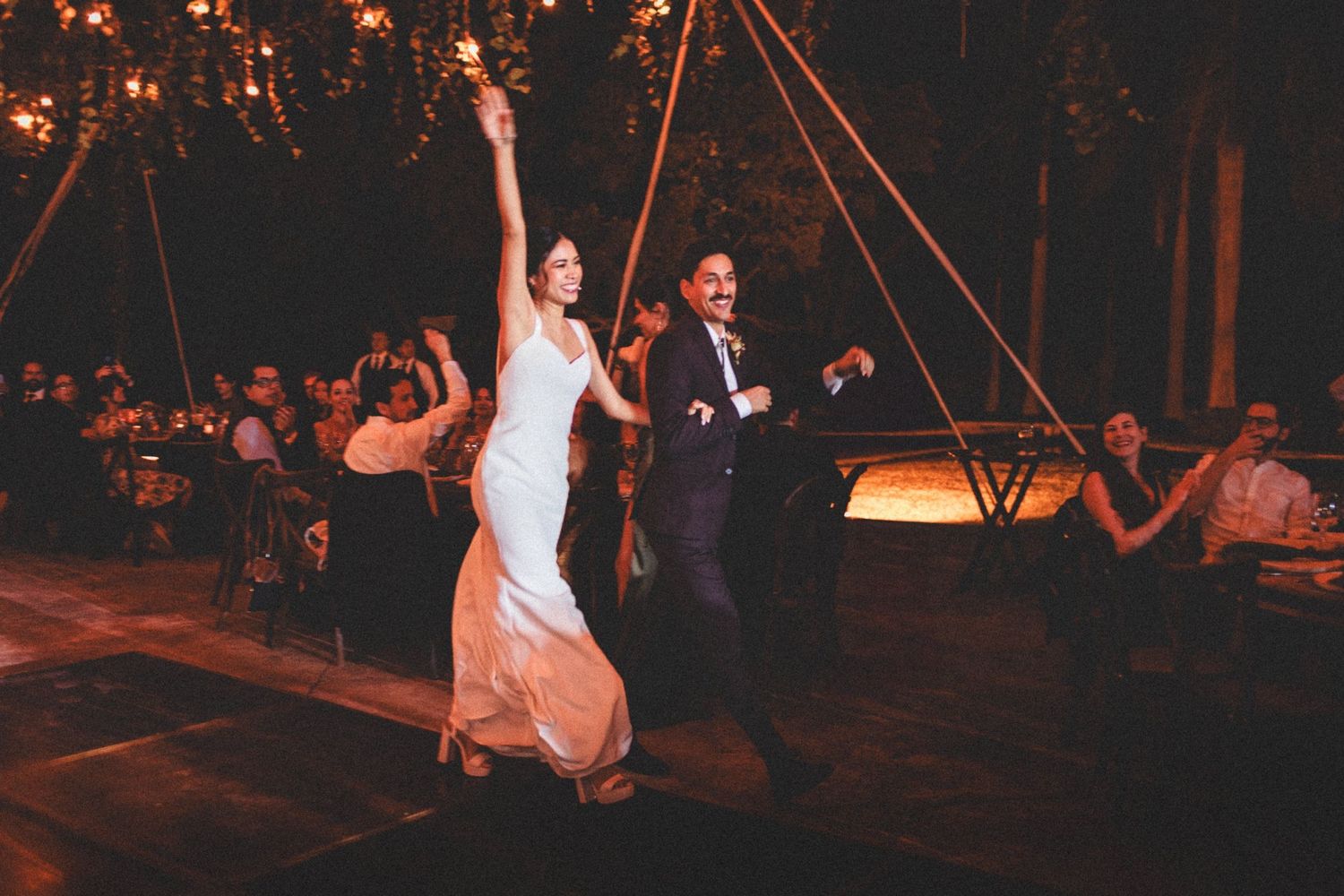 People celebrate at an outdoor evening wedding reception with string lights and decorative illumination creating a magical atmosphere.