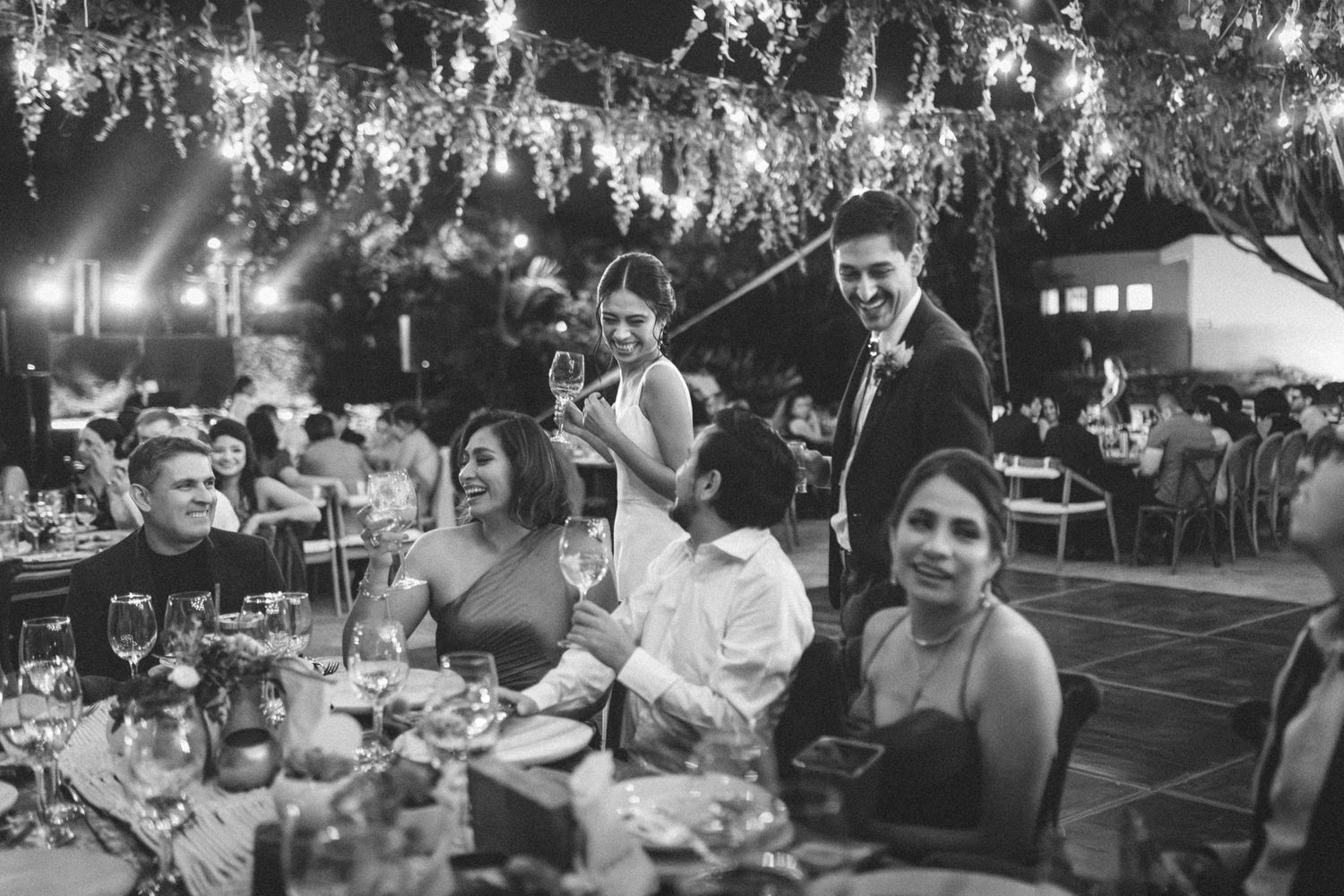 Black and white photo of guests enjoying an elegant outdoor evening reception with illuminated trees and dining tables.