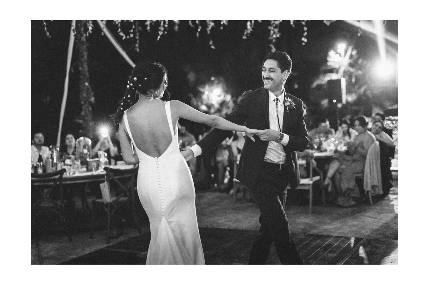 A couple shares a romantic dance at their wedding reception as guests look on from tables at an outdoor venue.