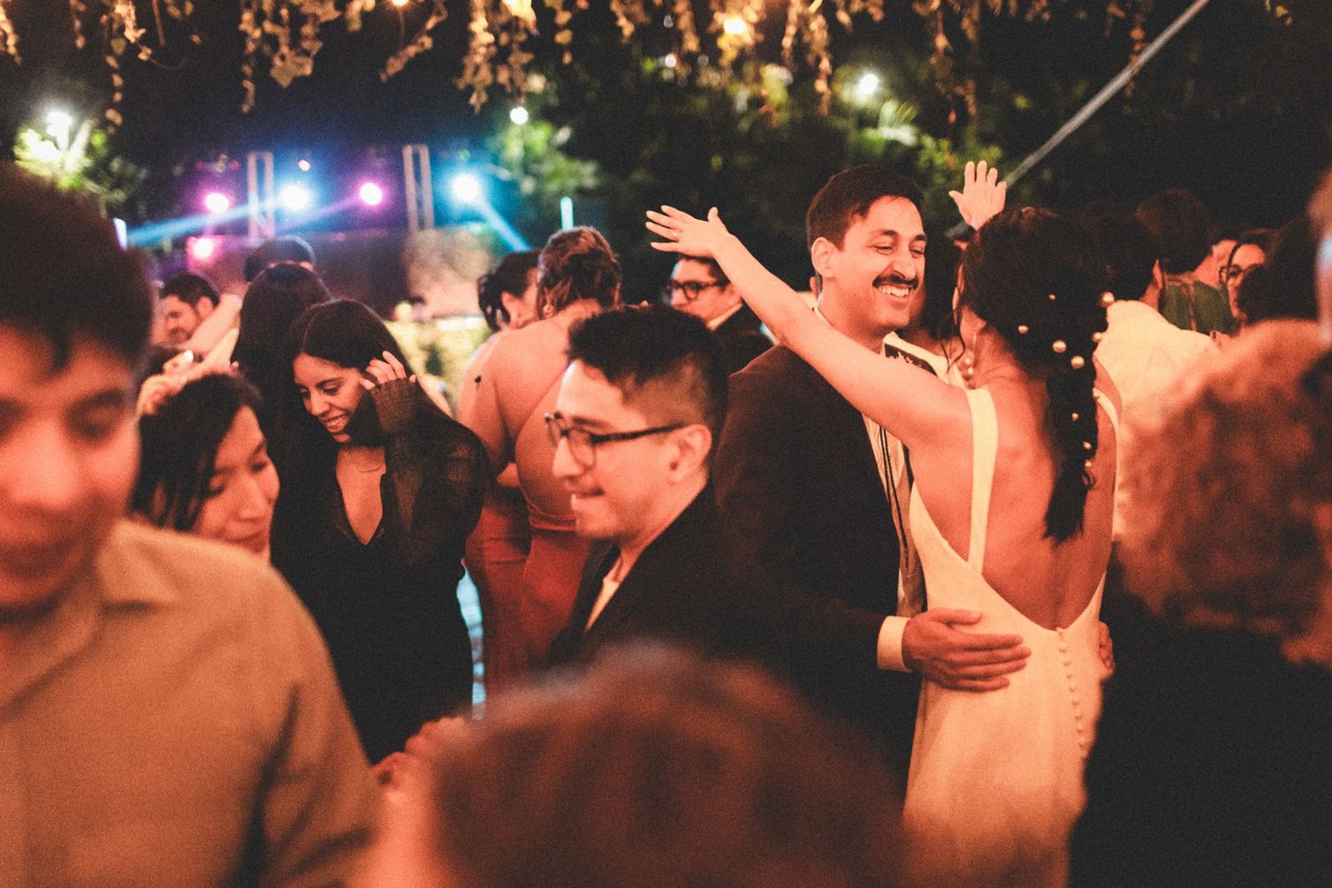 A crowd of people dancing and celebrating at a nighttime outdoor party with glowing lights in the background.