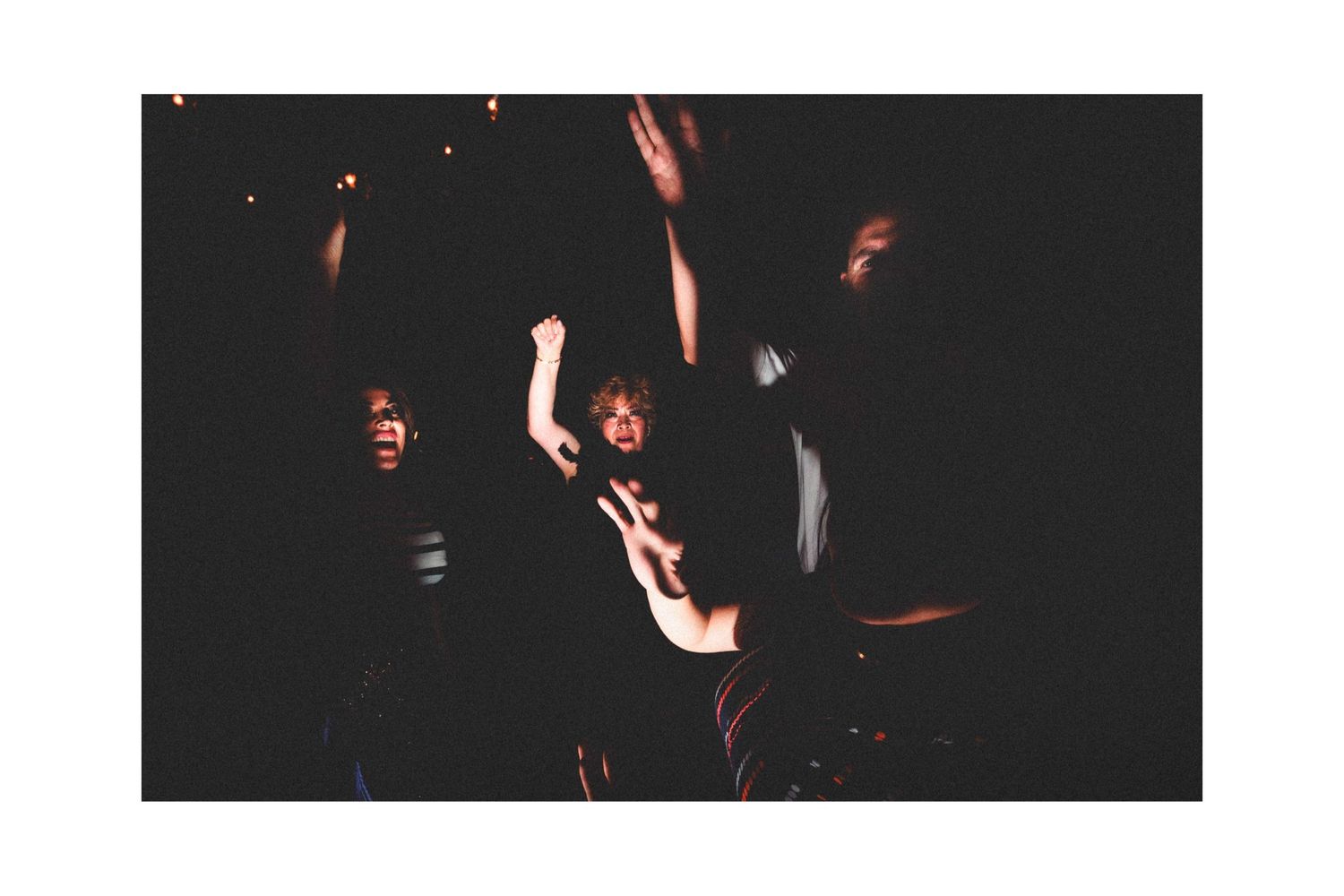 Dark concert scene with silhouetted figures raising hands against a dim red-lit backdrop.