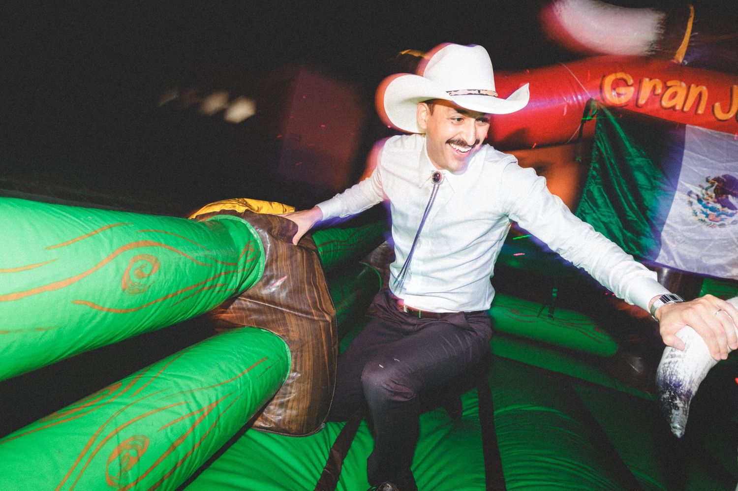 Mechanical bull rider attempts to stay mounted on red and black bull in dimly lit bar with green lighting.