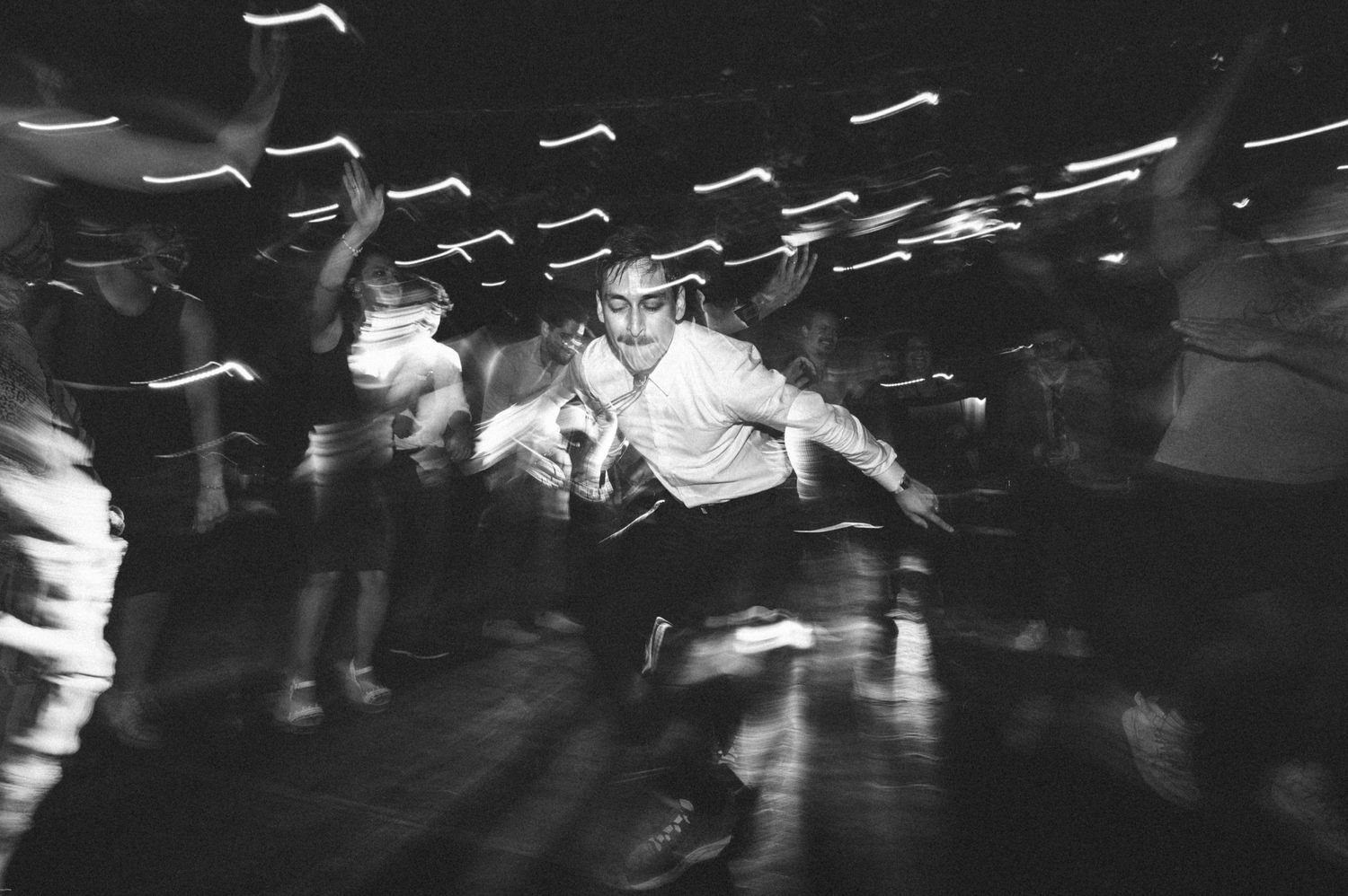 Black and white photograph of people dancing energetically at a nightclub with motion blur and streaking lights.