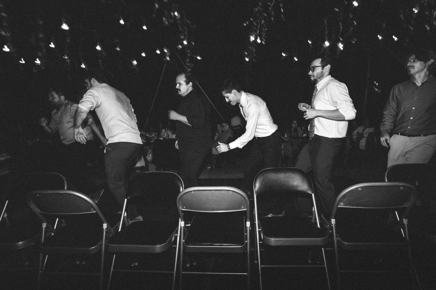 A black and white photograph shows people dancing and moving among metal folding chairs at an event.