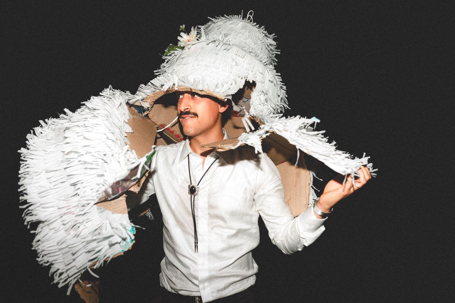 Performer in white outfit poses dramatically with large white feather boas against dark background.
