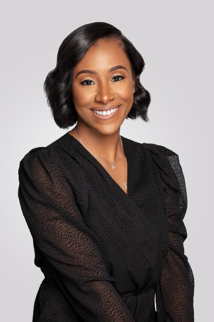 Professional headshot of woman in black sheer blouse smiling warmly against gray background.