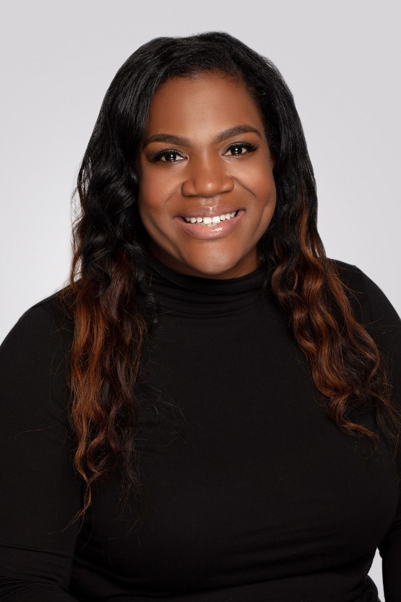 Professional headshot of woman in black turtleneck with ombre wavy hair against gray background.