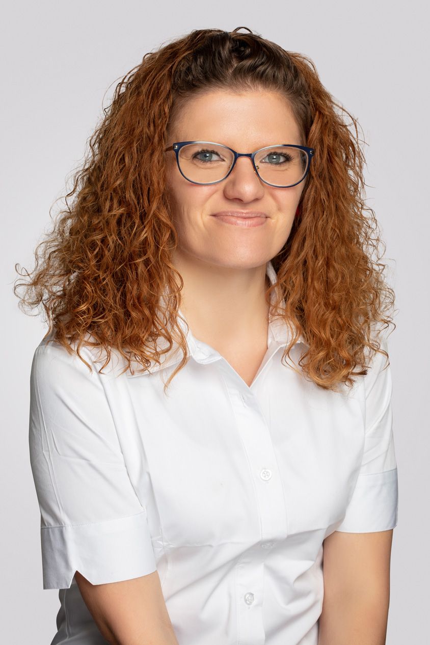 Professional headshot of woman with curly red hair wearing glasses and white shirt.