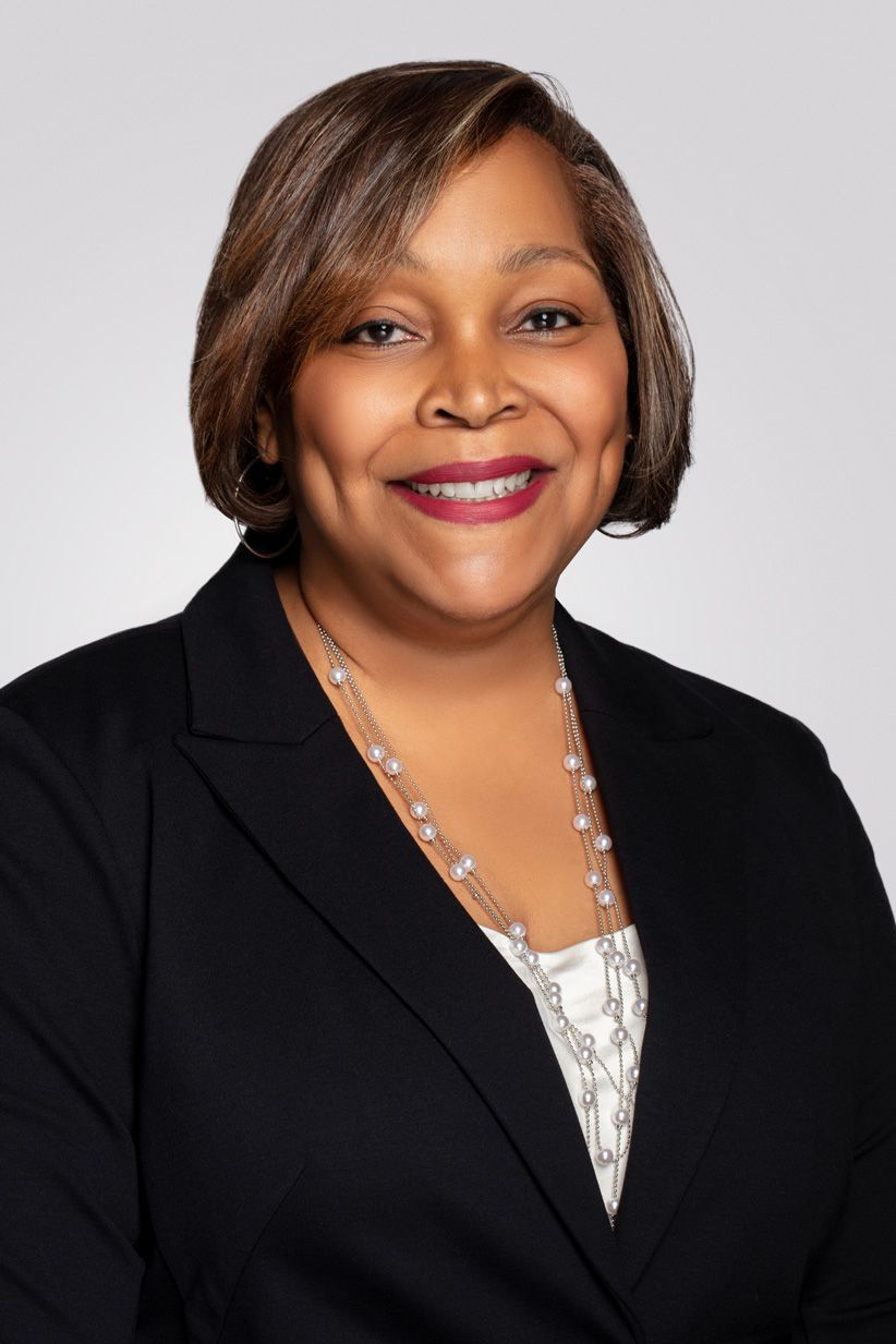Professional headshot of woman in black blazer and patterned top with layered necklace.