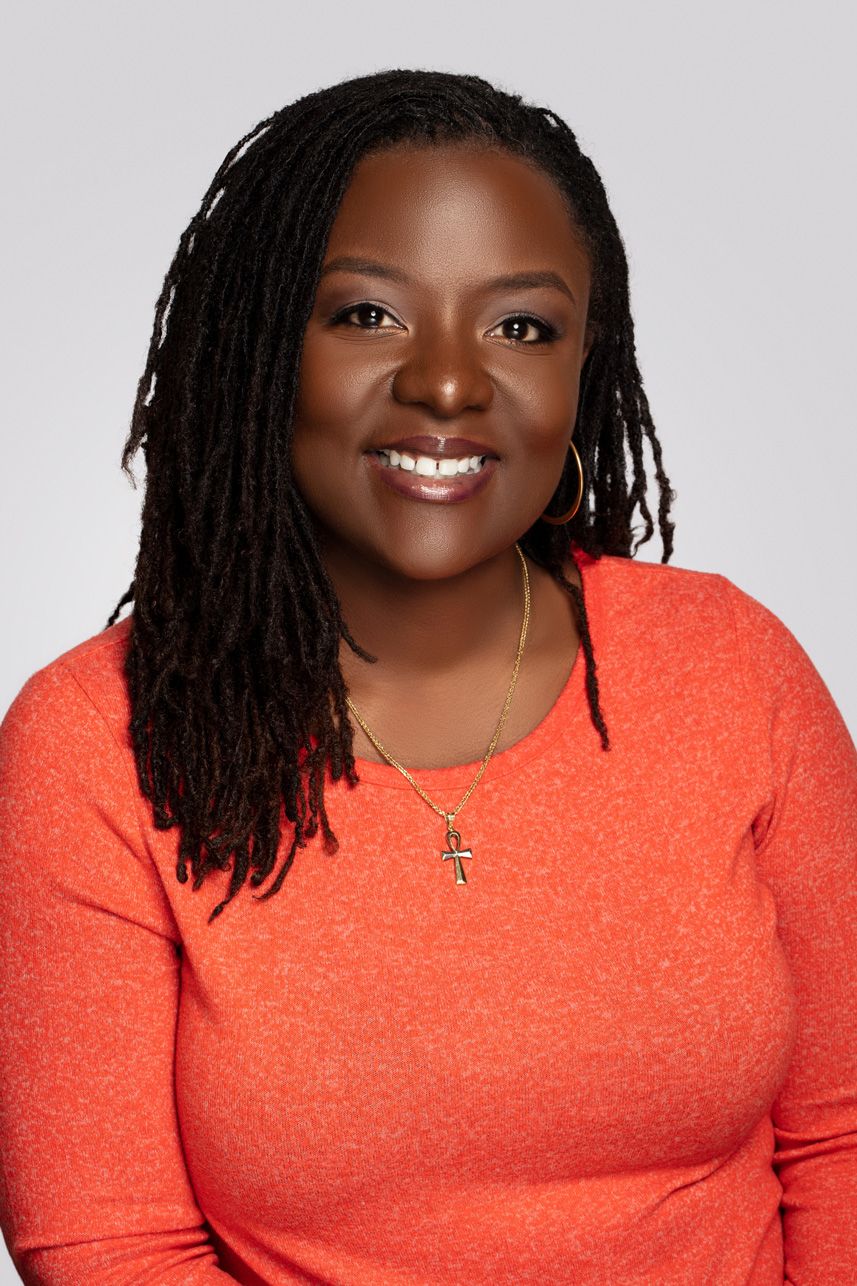 Warm studio portrait featuring someone in a bright coral sweater with long locs and cross necklace.
