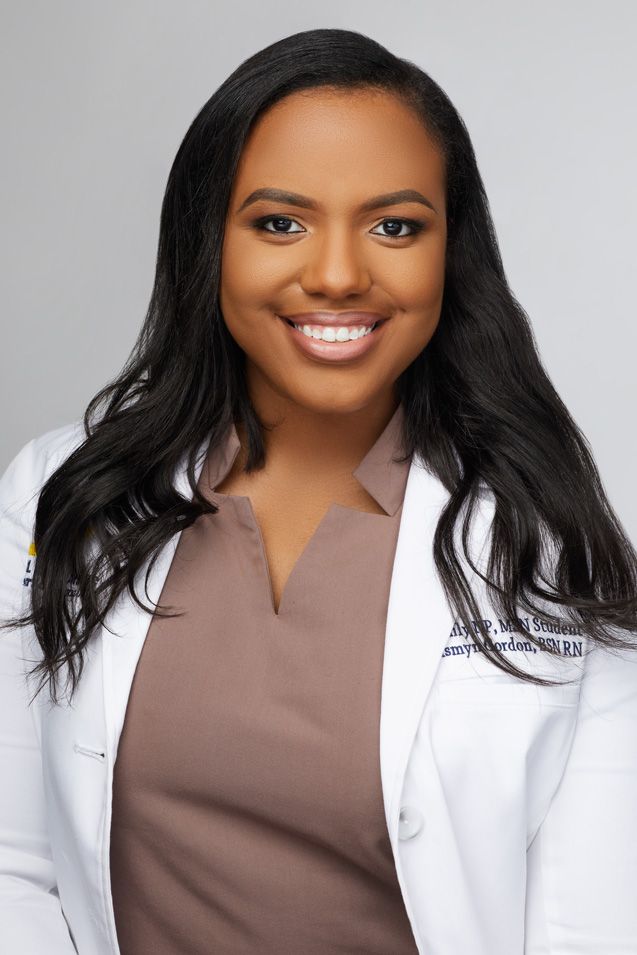 Professional headshot of a healthcare worker wearing a white lab coat and brown scrubs with a bright smile against gray background.