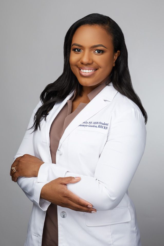 Professional healthcare provider wearing a white lab coat and brown top poses confidently against grey background.