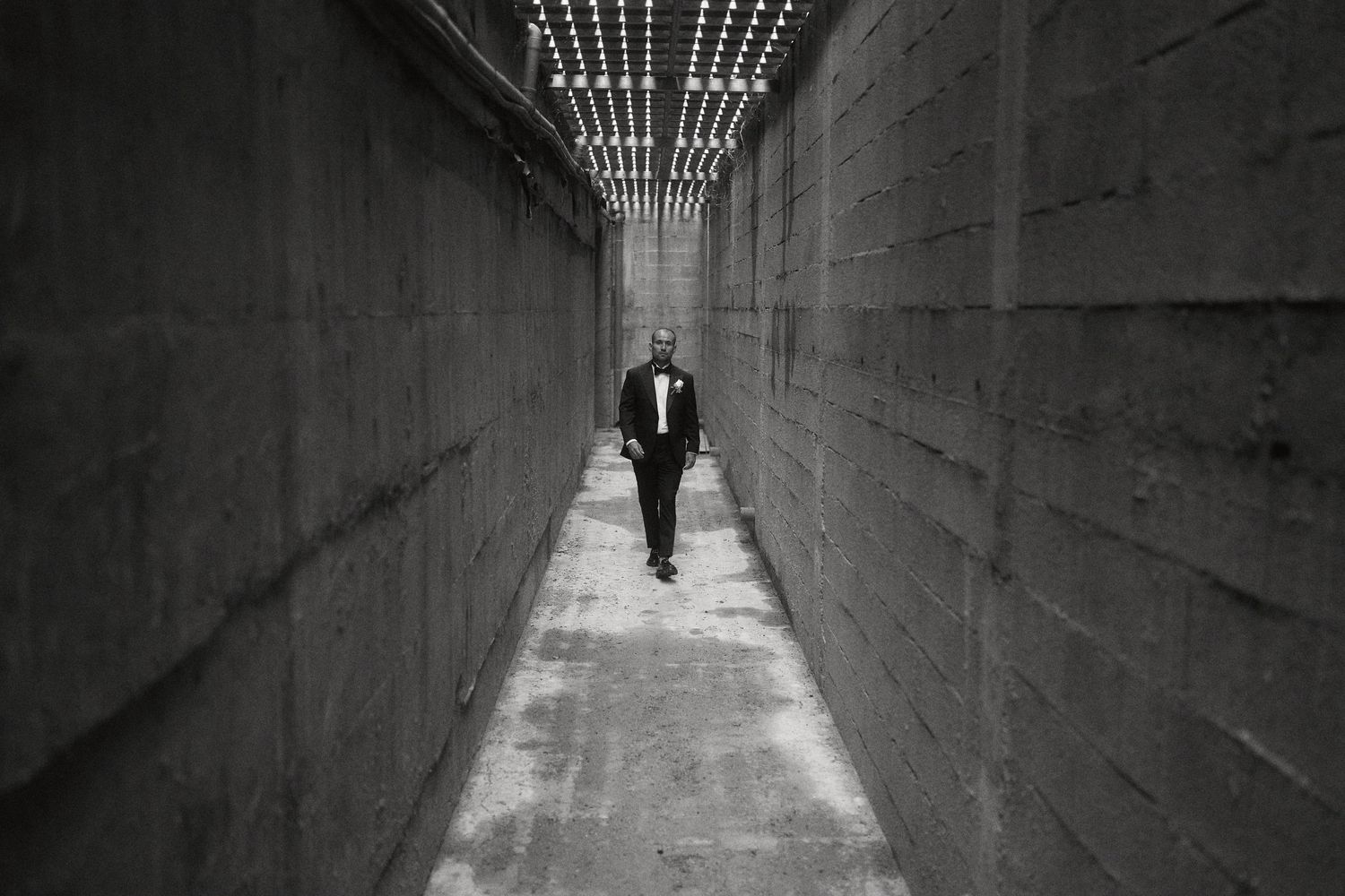 A black and white photo of a figure walking through a narrow concrete corridor with dramatic lighting from above.