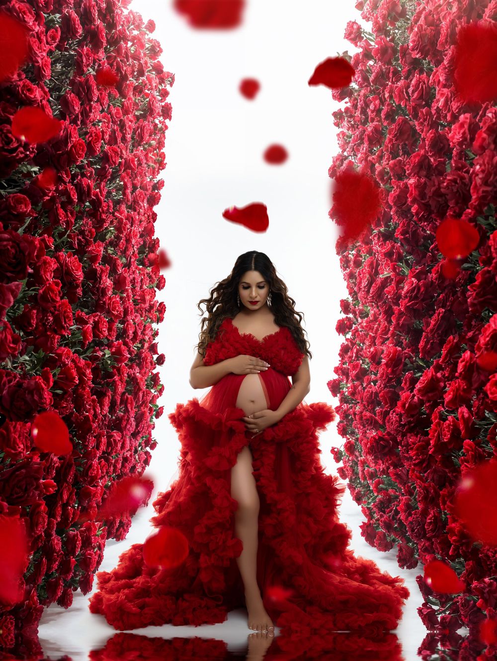 A figure in a flowing red dress poses among red rose walls in a romantic maternity photoshoot setting.