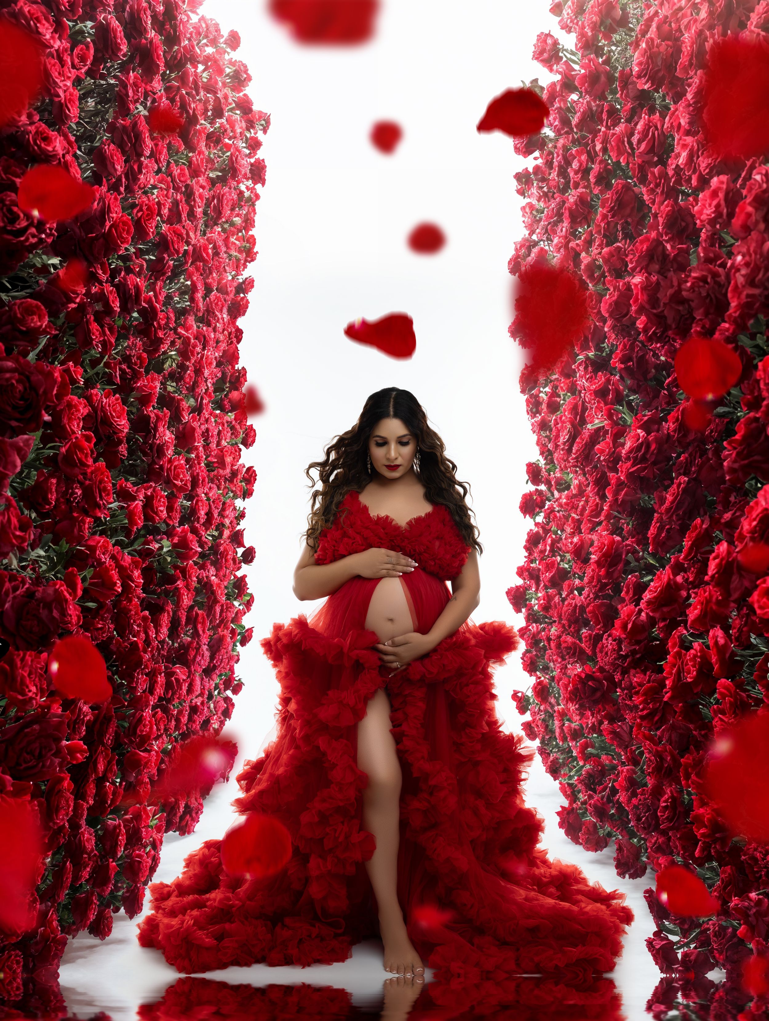 A figure in a flowing red dress poses among red rose walls in a romantic maternity photoshoot setting.