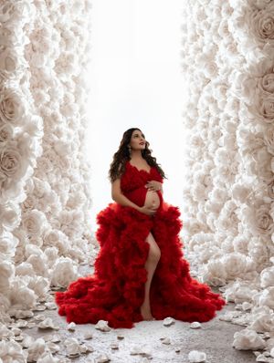 Ethereal maternity photo with a flowing red tulle gown against white floral backdrop in dramatic lighting.