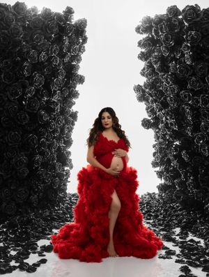 Dramatic maternity portrait featuring figure in flowing red gown against black and white floral backdrop.