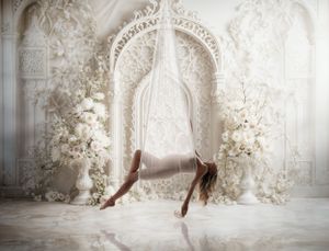 A figure in ballet pointe shoes performs artistic dance movements in an elegant white ornate room with floral decor.
