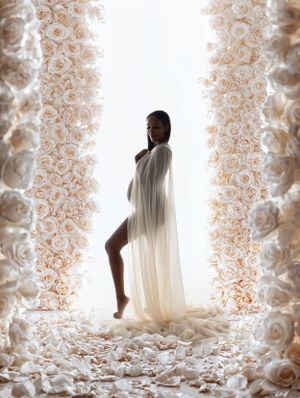 Artistic maternity photo with flowing white fabric between rose-adorned columns against bright background.