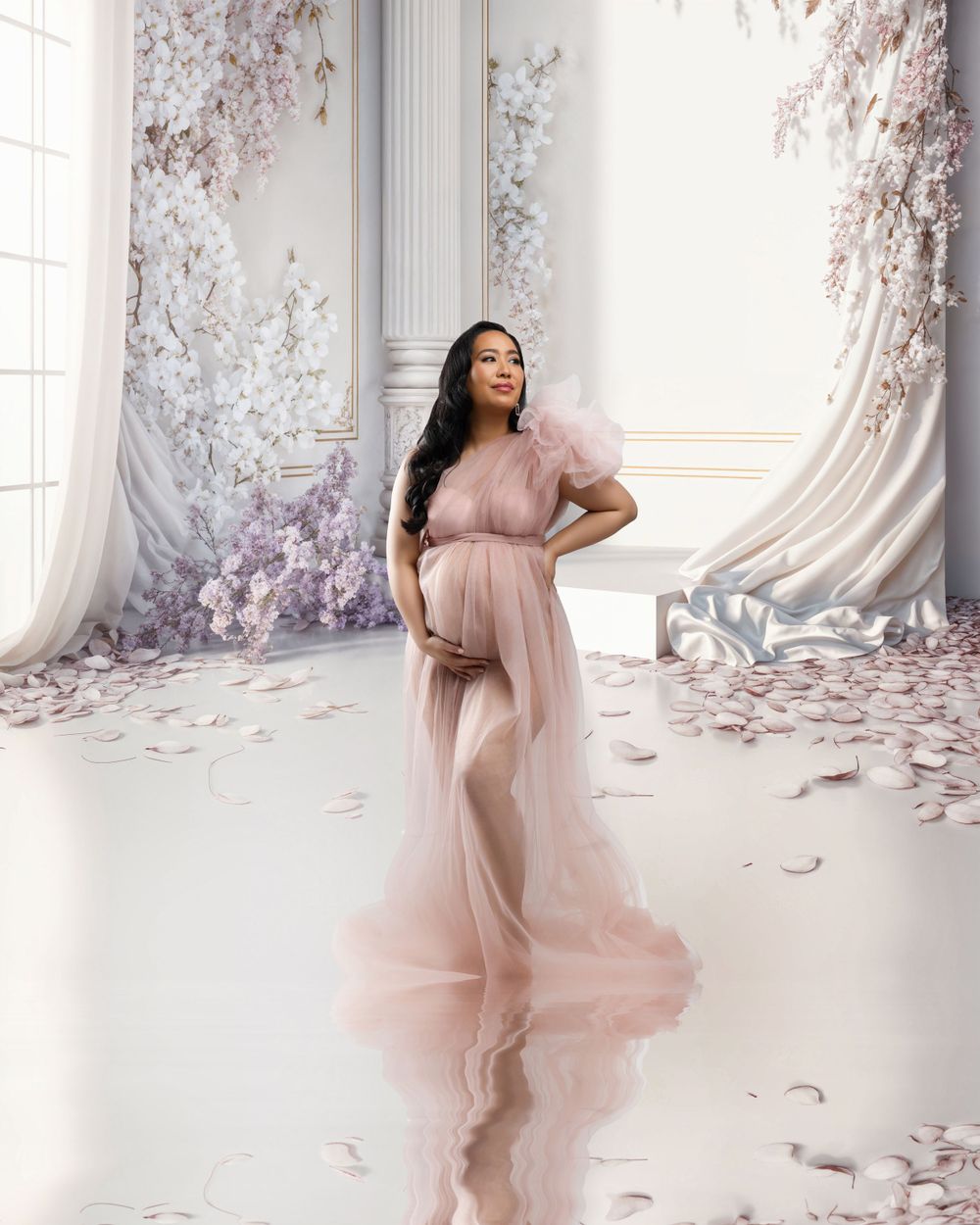 Ethereal maternity portrait in pink flowing gown with cherry blossoms in elegant white room setting.