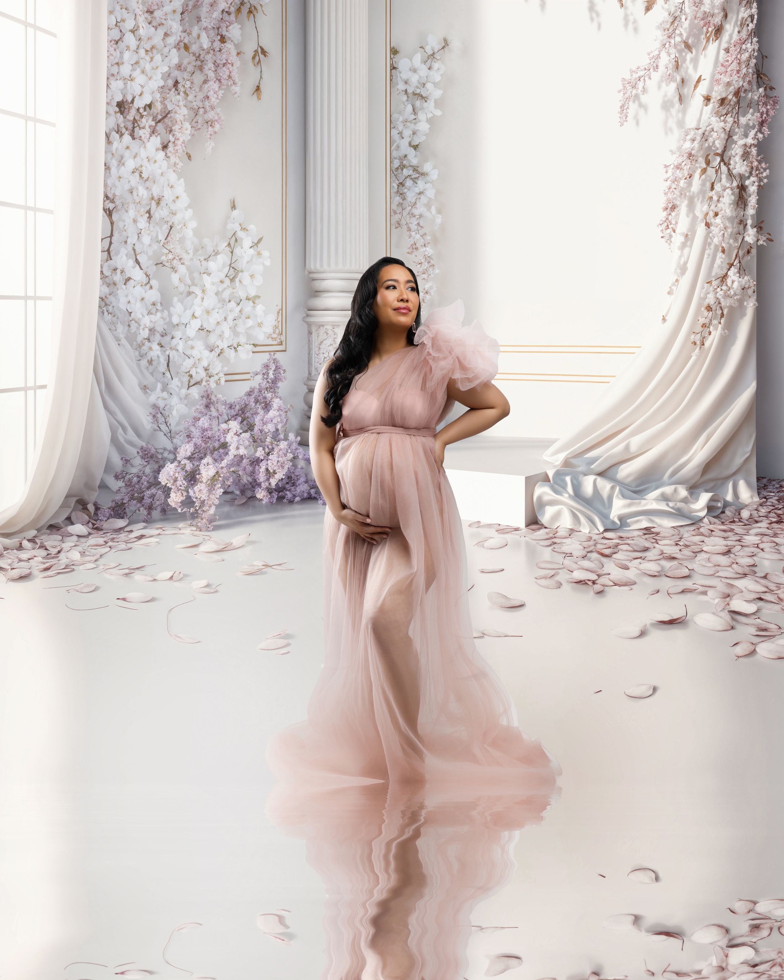 Ethereal maternity portrait in pink flowing gown with cherry blossoms in elegant white room setting.