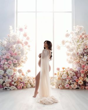 Ethereal portrait of someone in a sheer white dress standing between two luxurious floral arrangements by a bright window.
