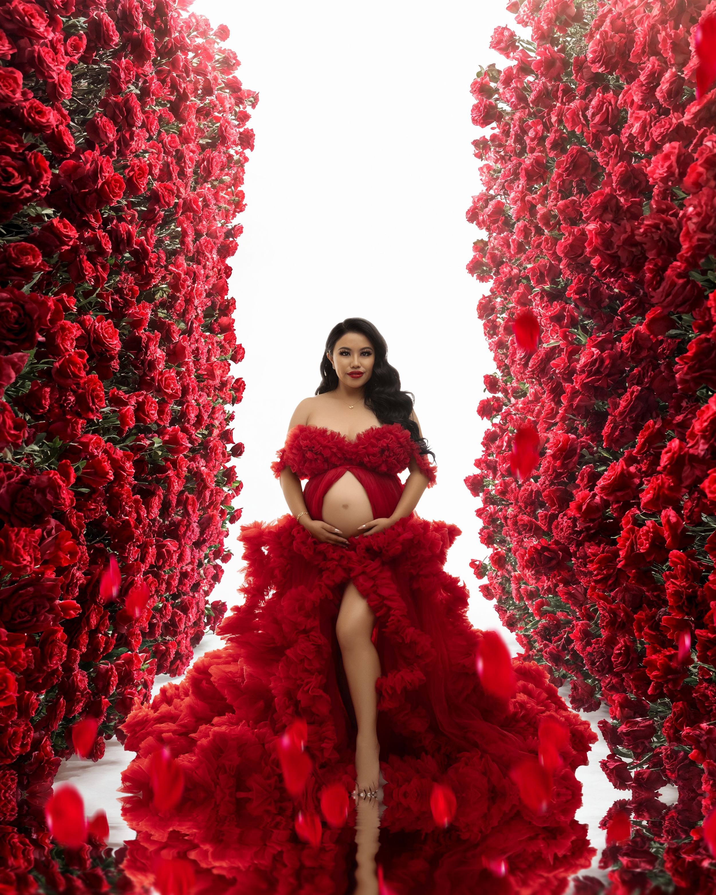 Dramatic maternity photo with a flowing red gown and tulle dress surrounded by vibrant red rose walls.