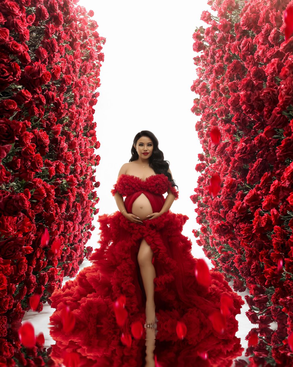 Dramatic maternity photo with a flowing red gown and tulle dress surrounded by vibrant red rose walls.