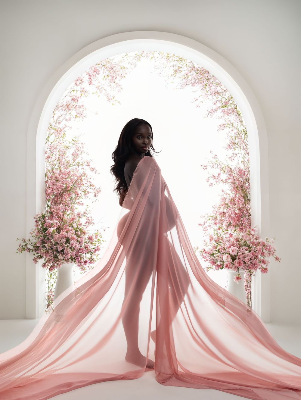 Silhouette stands in arched doorway framed by pink cherry blossoms wearing flowing pink maternity gown in ethereal light.