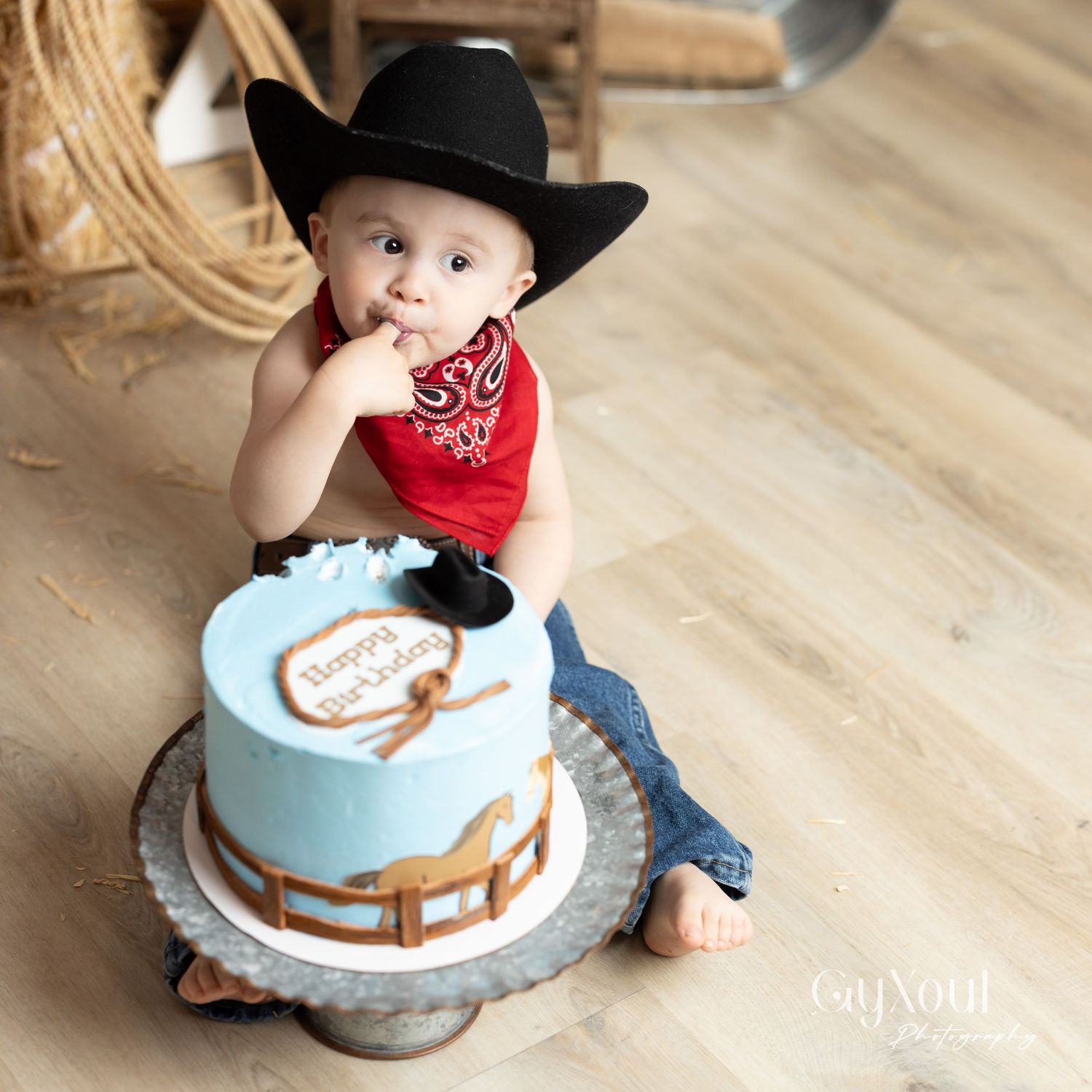 Baby in cowboy attire with a black hat and red bandana sits next to a light blue cake with brown decorative elements.