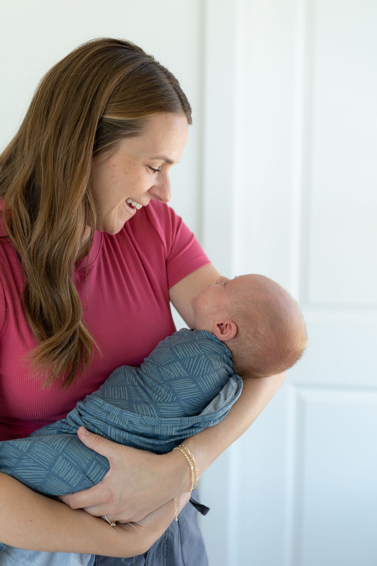a mom in a bright pink shirt smiling at her newborn baby boy swaddled in a blue blanket