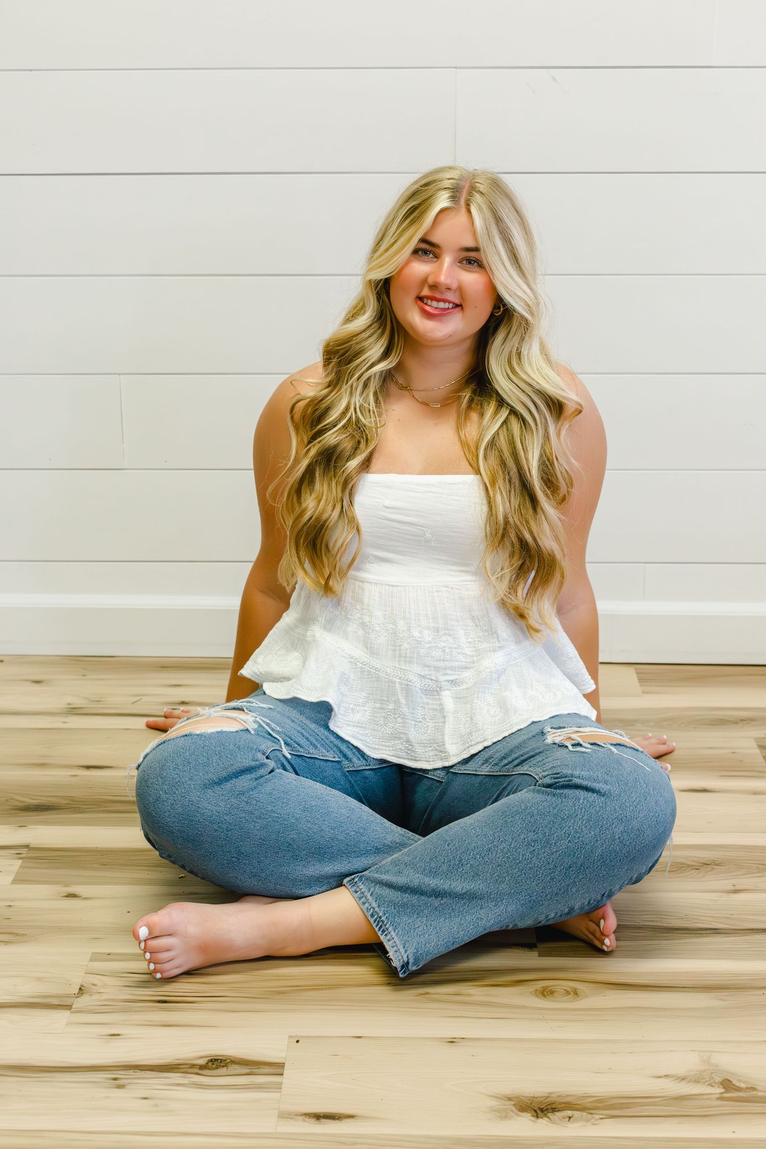 Casual indoor portrait with long wavy blonde hair wearing white top and jeans sitting cross-legged on wooden floor.