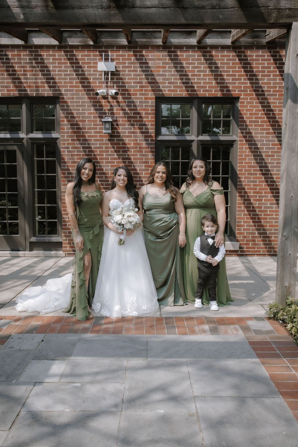 Bridal party in green bridesmaid dresses and white wedding gown poses in front of brick building.