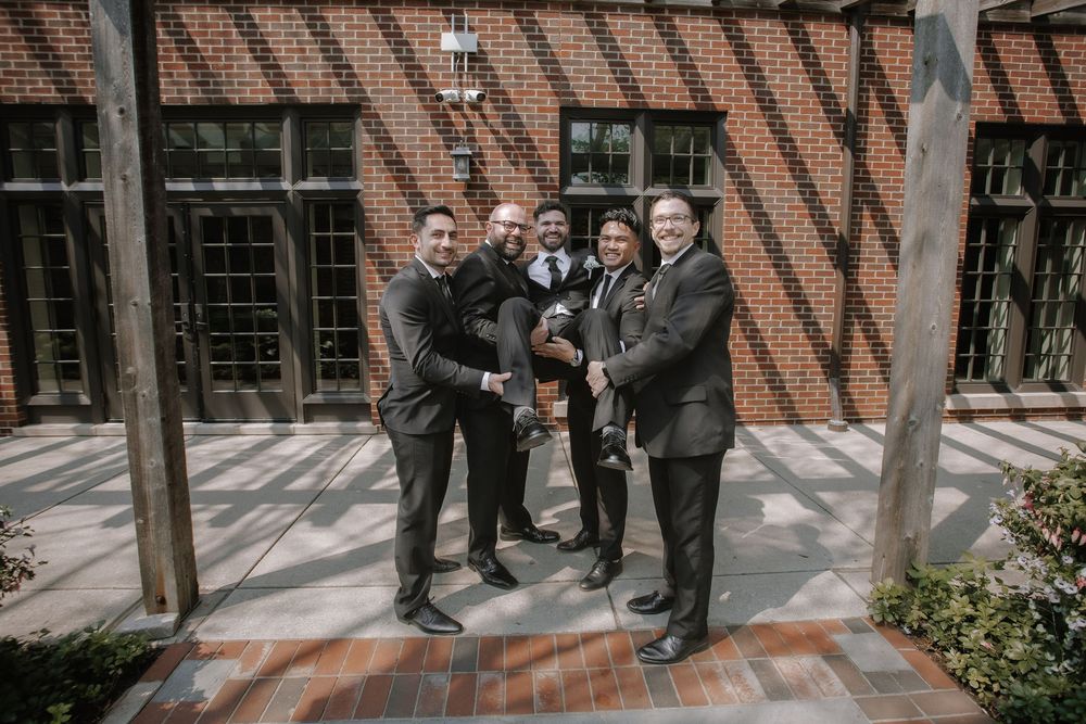 A group in matching dark suits poses together on a brick patio with shadow patterns from overhead beams.