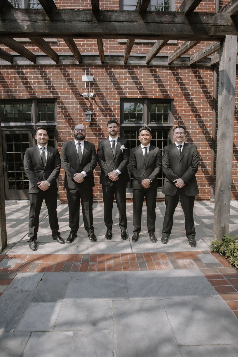 A line of groomsmen in matching dark suits and ties pose against a rustic brick wall at a wedding venue.