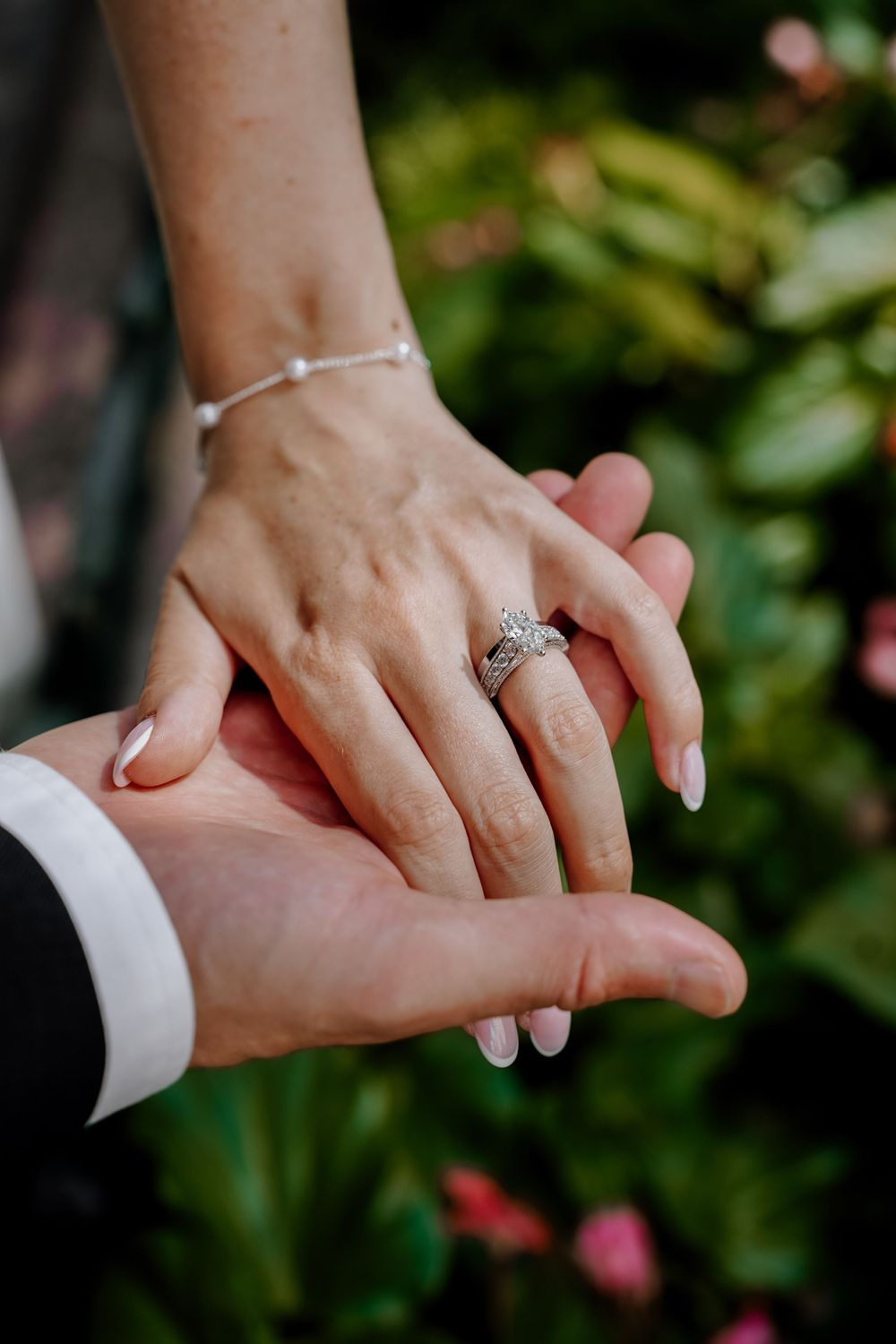 Close-up of newly engaged couple's hands with sparkly engagement ring and delicate bracelet against green garden background.