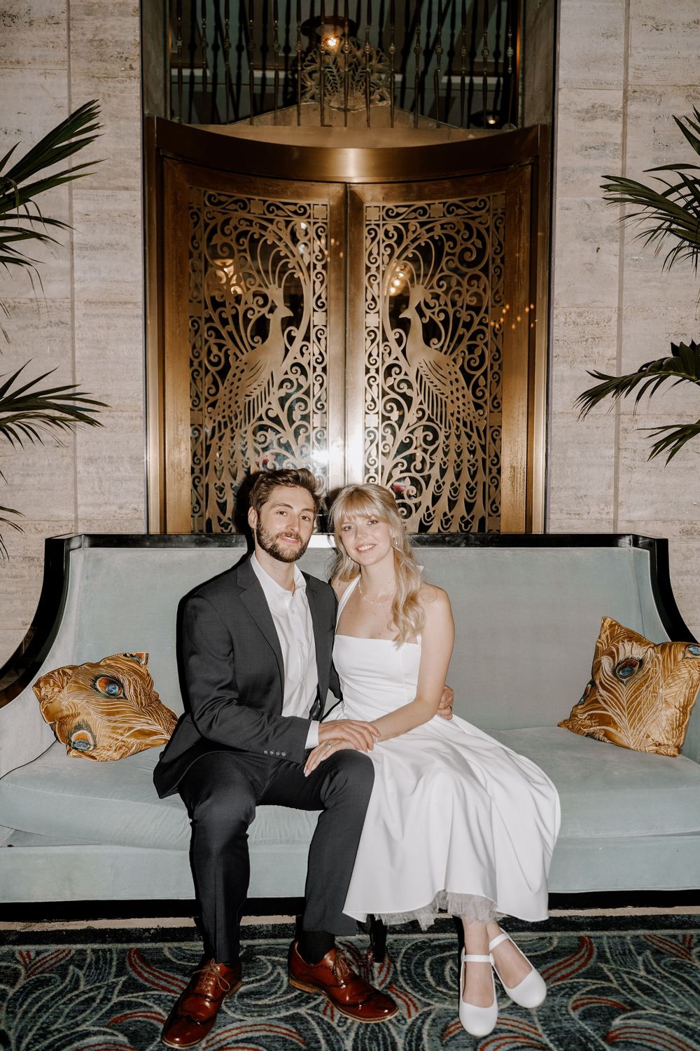 A couple sitting together on an elegant sofa with ornate decor and luxurious pillows in a vintage-style setting.