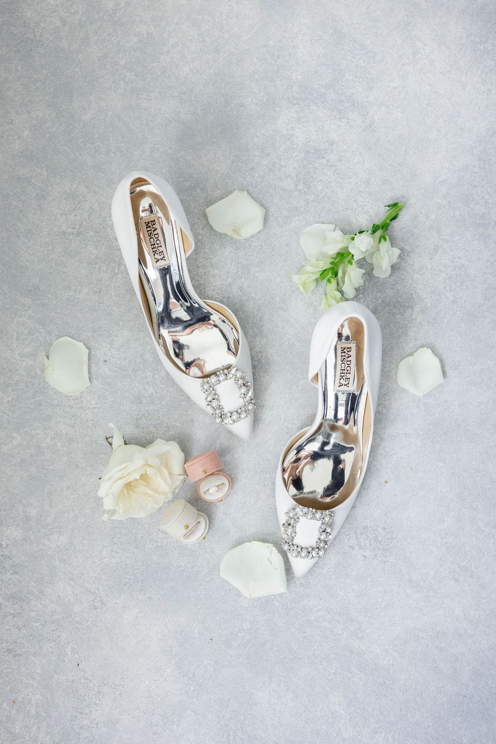 Elegant silver wedding shoes with crystal embellishments displayed with white rose petals and flowers on gray background.