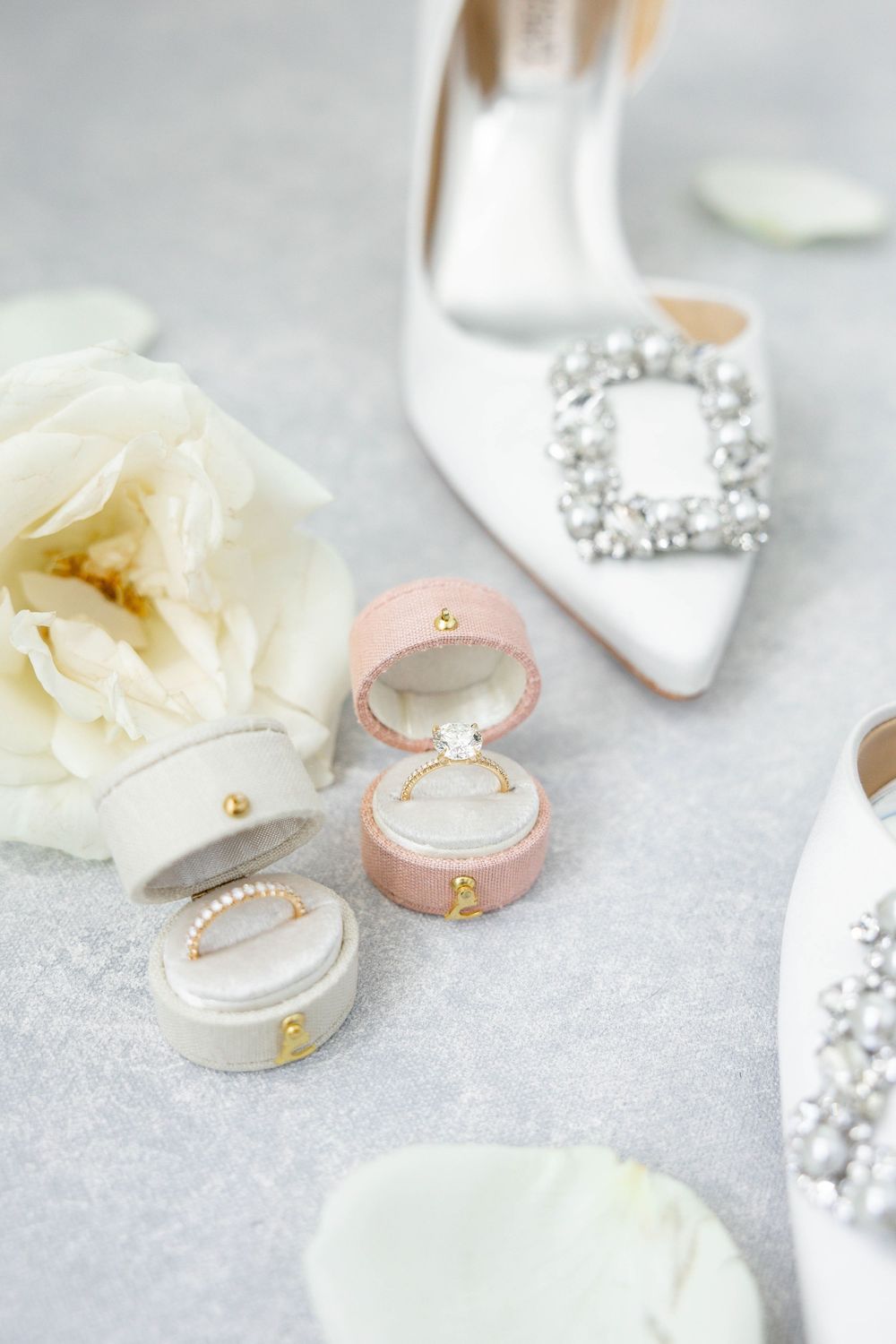 Elegant white bridal shoes with crystal embellishments displayed alongside delicate ring boxes and white rose petals.