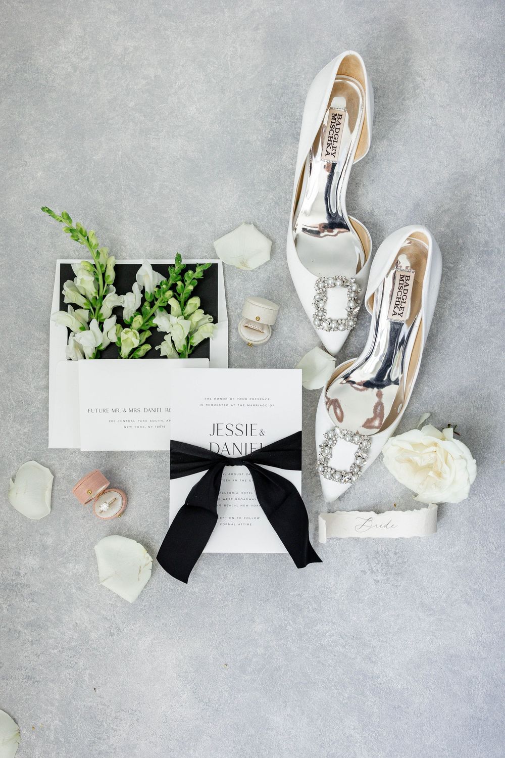 Elegant wedding invitations with white flowers, ribbon accents, and bridal shoes arranged on a light gray background.