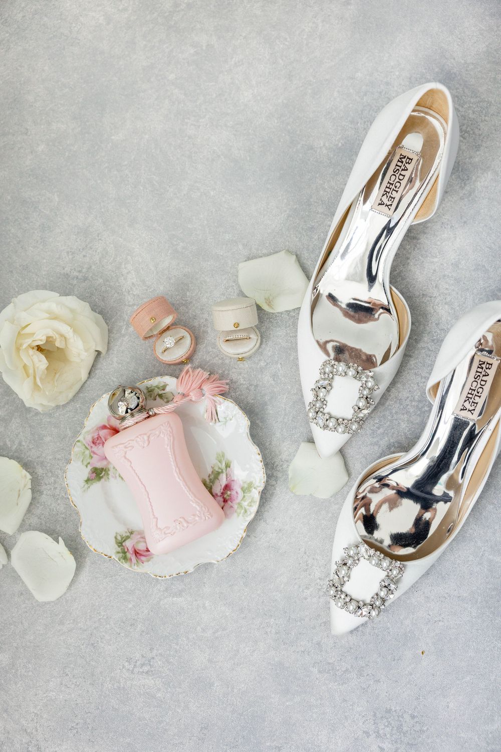 Elegant white wedding heels with crystal buckles displayed alongside pink ring box and delicate white rose petals.