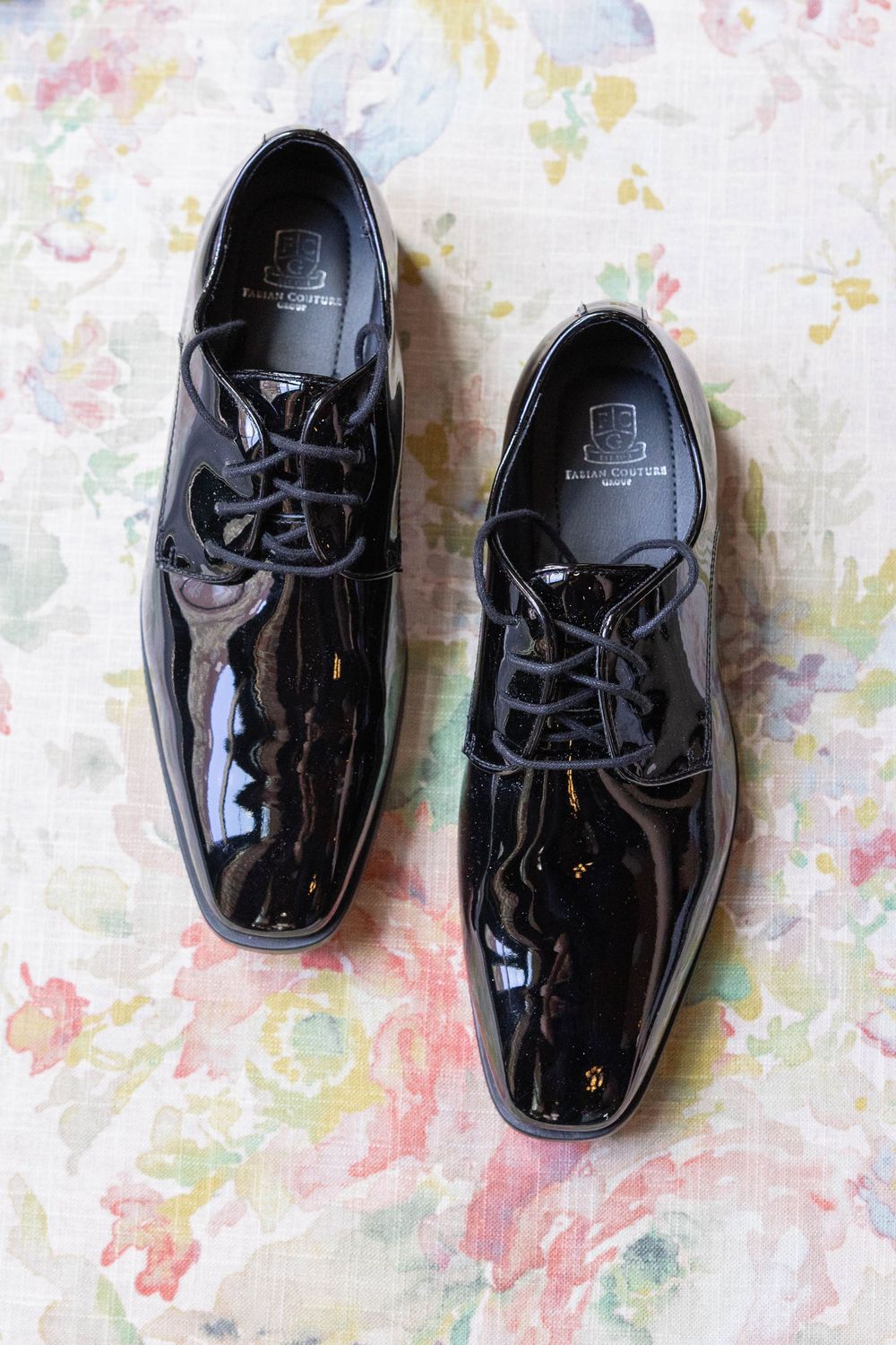 A series of three identical pairs of shiny black patent leather dress shoes arranged on a floral background surface.