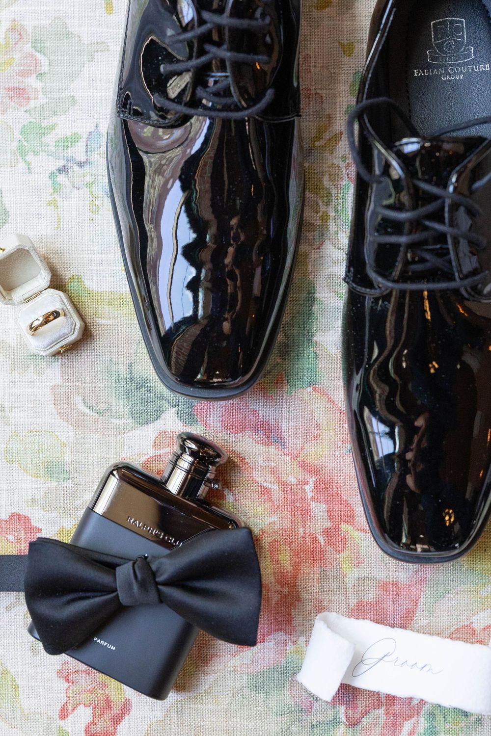 Black patent leather dress shoes with bow tie and cologne bottle arranged on colorful floral patterned surface.