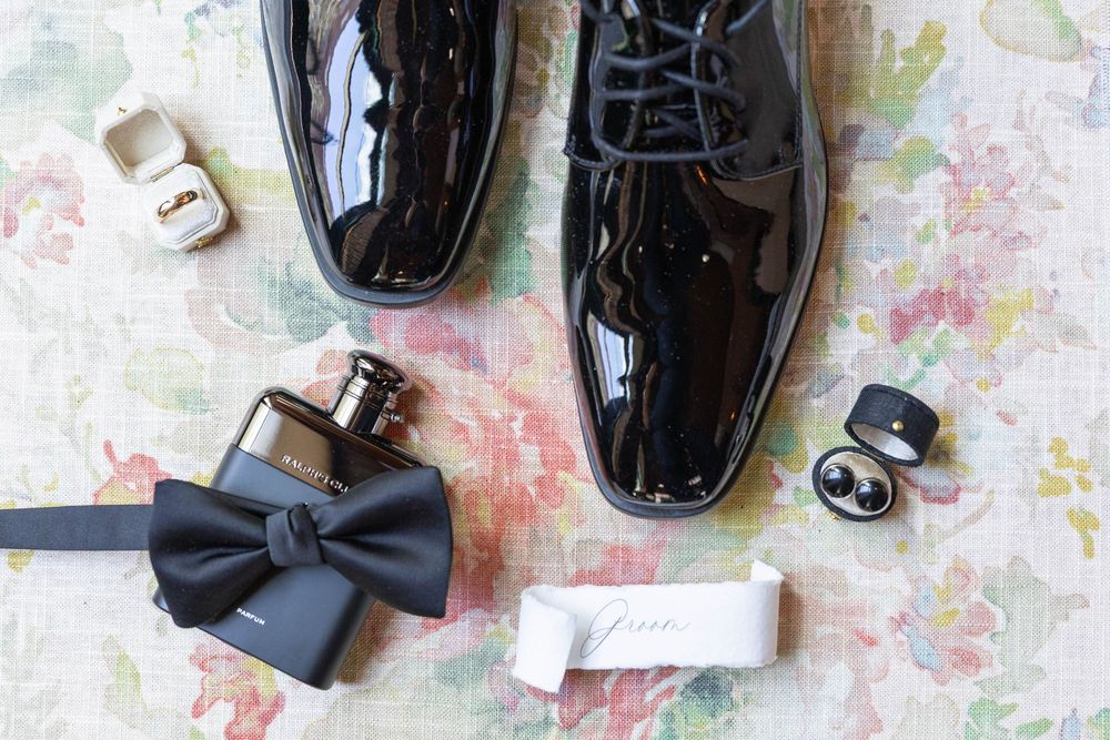 Black patent leather formal dress shoes with bow tie and cufflinks arranged on floral patterned fabric background.