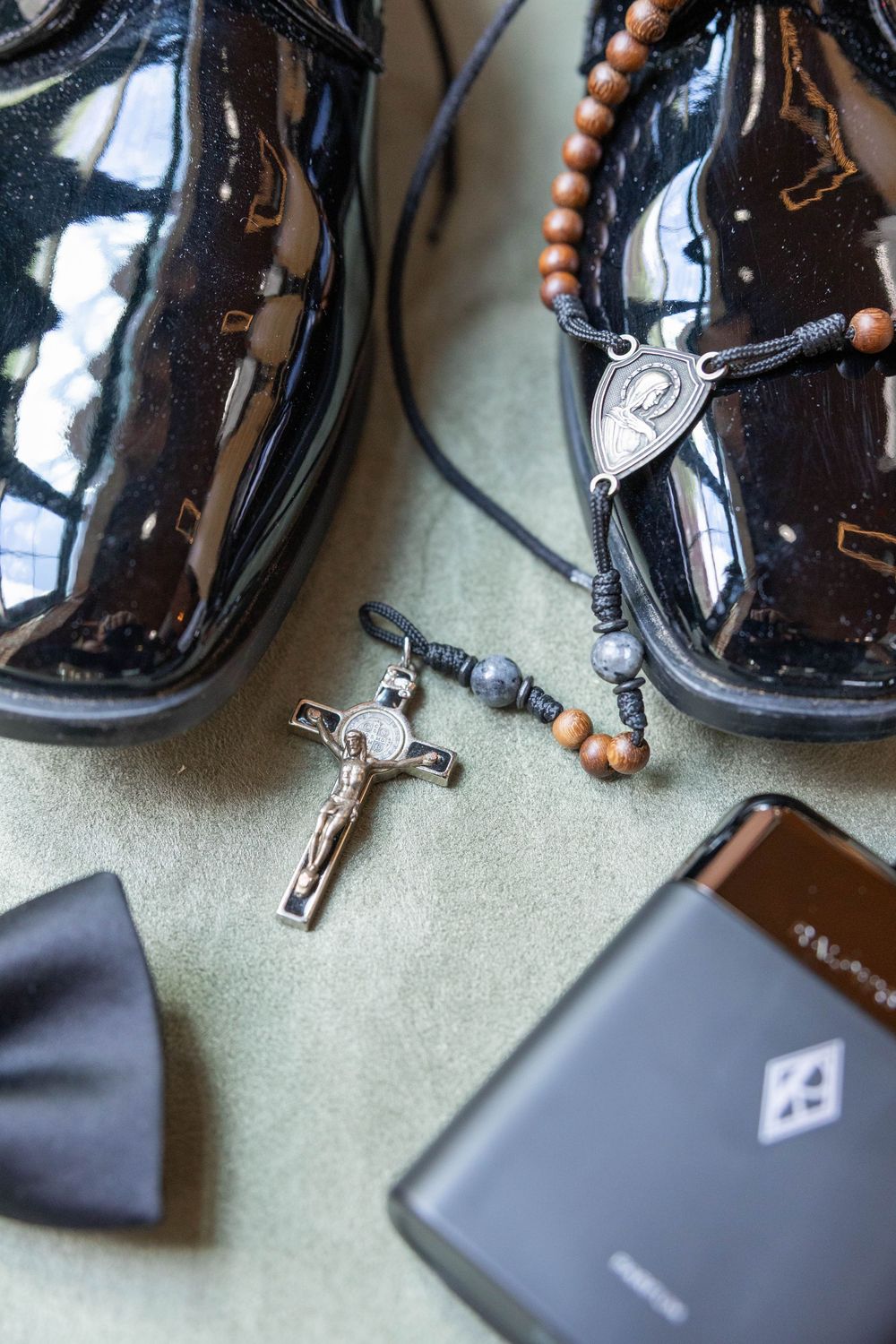 Shiny dress shoes with rosary beads, crucifix, black bow tie and wallet arranged on green background.